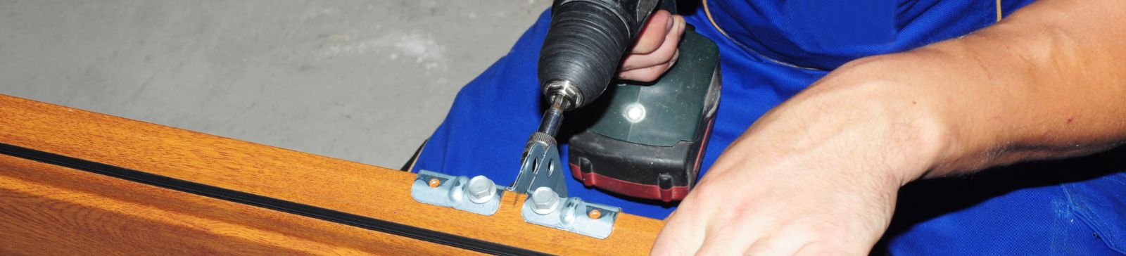 Close-up of a professional technician in blue overalls using a power drill to install a hinge on a wooden garage door panel during a project in Fern Park.